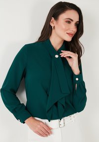 Teal silk blouse with a tied front detail, button accents on sleeves, and a smooth texture, paired with a white belt and fitted pants.