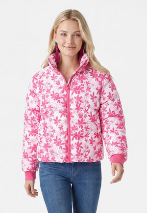 Junge Frau trägt eine rosa-weiß geblümte Steppjacke mit Jeans und langen Quastenohrringen, lächelnd und nach vorne blickend.