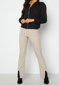 Giacca bomber nera a costine con chiusura zip, abbinata a pantaloni beige attillati con fessure laterali e stivaletti neri alla caviglia.