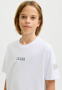 Valge puuvillane T-särk, millel on sinine "JACK & JONES" logo rinnal ja ruudukujuline kleebis vasakul varrukal. Lihtne ümmargune kaelus.