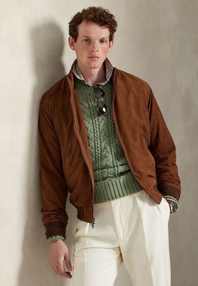 Tânăr cu păr creț purtând o jachetă maro din piele întoarsă peste un pulover verde cu model de torsadă și pantaloni crem, sprijinit de un perete bej.