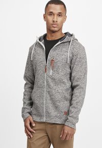 Gray zip-up hoodie made of textured fabric with a hood, featuring brown accents and a front pocket. Worn with a black shirt and khaki pants.