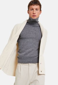 Cardigan en tricot ivoire sur un pull à col roulé gris, complété par un pantalon beige. Le cardigan présente une texture côtelée et des détails de boutons.