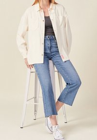Light cream oversized shirt in ribbed texture with two chest pockets, paired with mid-rise blue denim jeans featuring a cropped hem. White sneakers.