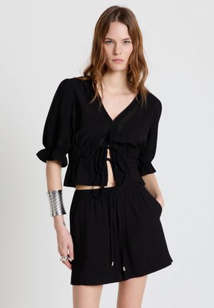 Jeune femme portant un chemisier court noir à manches bouffantes et un short noir assorti avec cordon de serrage, bracelet manchette argenté, debout devant un fond uni.