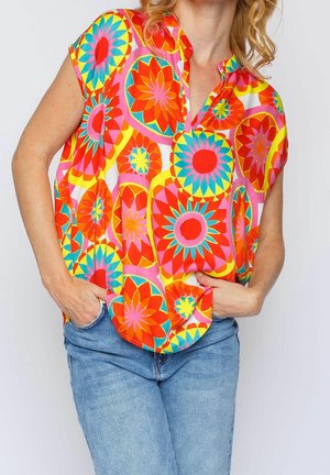 Donna che indossa un top senza maniche dai colori vivaci con grandi motivi floreali geometrici in rosso, arancione, giallo, rosa e blu, con jeans blu, mani nelle tasche.