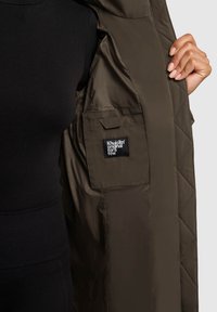 Kootud oliiviroheline jope, mille sisepinnal on must silt "Khujo™ original for a few" ning küljetasku detail. Sile tekstuur.