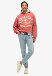 Sudadera coral de gran tamaño con impresión "Superdry Surf Contest", jeans de mezclilla azul claro y zapatillas beige. Estilo casual, ajuste relajado con un look en capas.