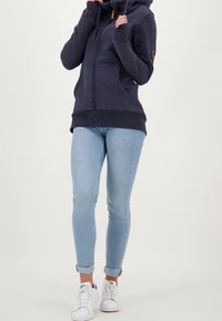Sudadera azul oscuro con cremallera, puños y dobladillo acanalados, con un cuello alto, cordón ajustable y un patrón de manchas rosas. Combinada con jeans azul claro.