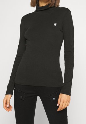 Femme portant un haut noir ajusté à col roulé avec un petit logo blanc sur la poitrine et un pantalon noir avec des poches à boutons.