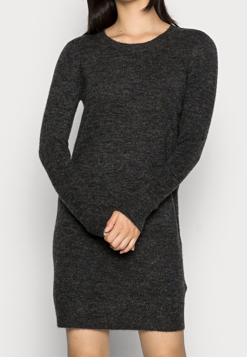 Femme portant une robe-pull en maille gris foncé à manches longues avec un col rond, debout, les mains jointes devant.