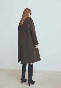 Abrigo marrón de cuadros con un corte holgado y abertura trasera, combinado con botas negras hasta la rodilla y un diseño elegante. Textura suave y forma entallada.