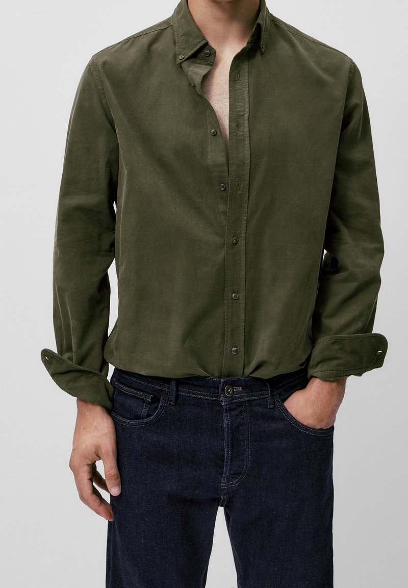 Homme portant une chemise kaki à boutons avec les manches retroussées et un jean bleu foncé, une main dans la poche et le torse partiellement visible.