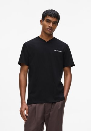 Mann mit kurzen lockigen Haaren, der ein schwarzes V-Ausschnitt-T-Shirt mit kleinem weißen "Karl Lagerfeld"-Text und lockeren braunen Hosen trägt.