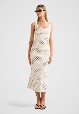 Νεαρή γυναίκα που φοράει εκρού midi φόρεμα με μαύρες πουά, μαύρα γυαλιά ηλίου και μαύρα σανδάλια, στέκεται μπροστά σε απλό φόντο.