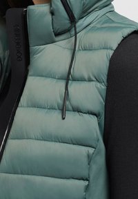Turkusowa pikowana kamizelka puchowa noszona na czarnej, prążkowanej bluzie z długim rękawem, z widocznymi czarnymi sznureczkami i zamkami błyskawicznymi.
