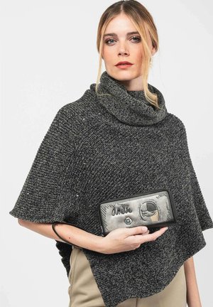 Mujer con cabello rubio que lleva un poncho de punto de color gris oscuro, sosteniendo una cartera gris y negra con costuras decorativas y una correa.