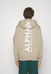 Bež hoodie s velikim bijelim logom "ALPHA INDUSTRIES" na leđima, kengur džepom i kapuljačom; ima rebraste manžetne i donji rub.