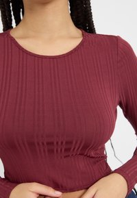 Bordo long-sleeve majica od rebrastog materijala s okruglim izrezom i naboranim detaljima na rubu. Teksturalni i priljubljeni dizajn.
