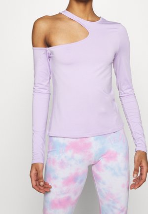 Kvinde iført en langærmet lavendelfarvet top med asymmetrisk udskæring ved skulderen samt pastelfarvede lyserøde og blå tie-dye leggings.