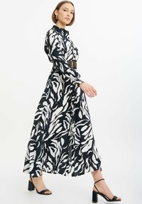 Schwarz-weißer Tierdruck-Langrock mit einem Kragen-Ausschnitt, Knopfverschluss und einem breiten schwarzen Gürtel. Das Modell trägt schwarze Blockabsatzsandalen.