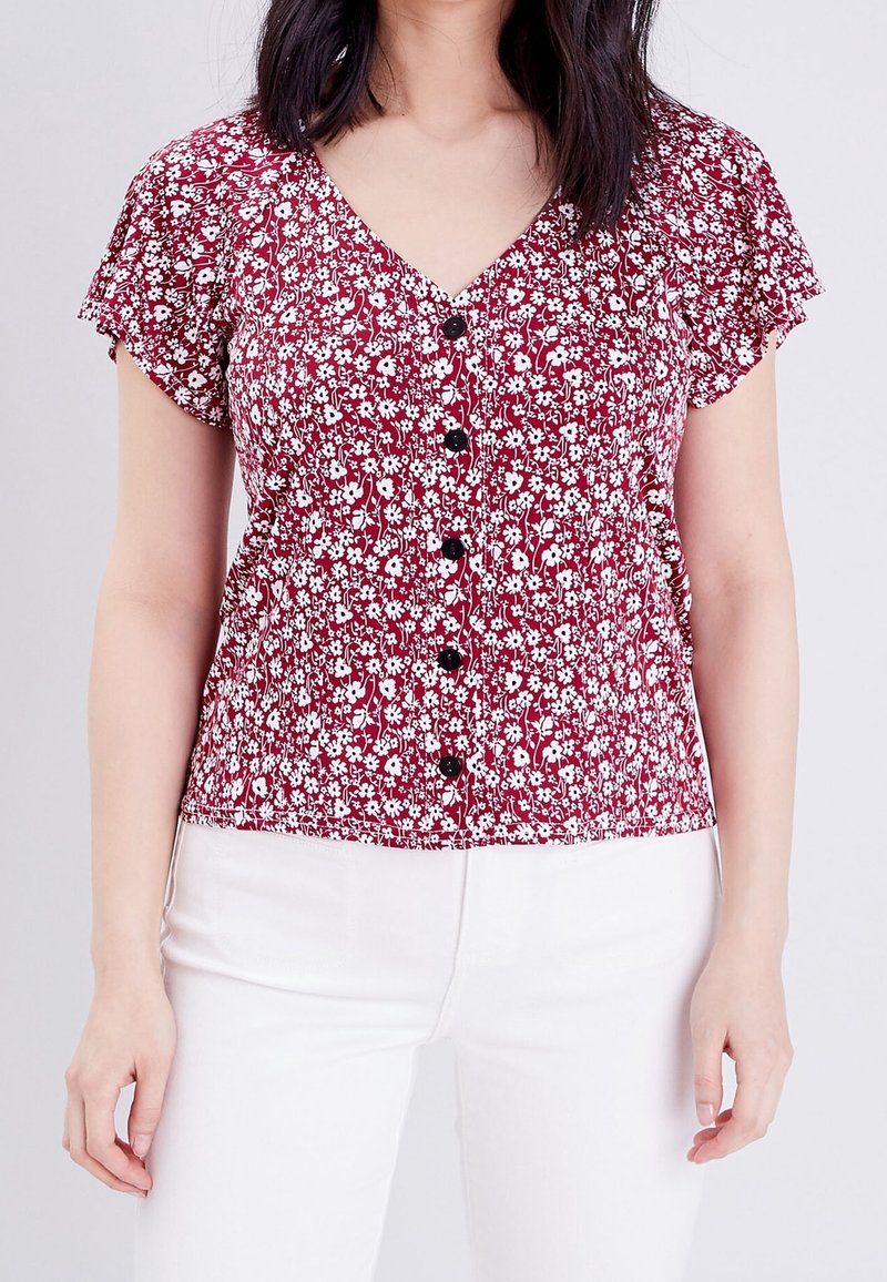 Blouse florale rouge avec un col en V, des manches courtes flottantes et des boutons noirs sur le devant. Présente une taille cintrée et un motif floral blanc.