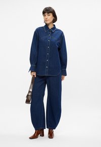 Σκούρο μπλε τζιν oversized πουκάμισο με κουμπιά μπροστά, μακριά μανίκια και αντίθετες ραφές, σε συνδυασμό με φαρδιά τζιν παντελόνια. Καφέ τσάντα.