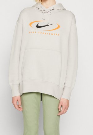 Person trägt einen hellbeigen Nike Sportswear Hoodie mit orange-schwarzem Logo, kombiniert mit hellgrünen, gerippten Hosen vor einem schlichten Hintergrund.