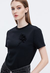 Camiseta de algodón negra con mangas cortas, que presenta un aplique floral negro texturizado con cuentas en el lado izquierdo.