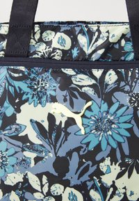 Floral-patterned tote bag in navy, light blue, and white. Features a textured material and a light green Puma logo. Durable handles.