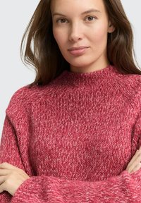 Junge Frau mit heller Haut und braunen Haaren, die einen dicken, strukturierten roten Strickpullover trägt, mit verschränkten Armen, nach vorne blickend mit neutralem Gesichtsausdruck.