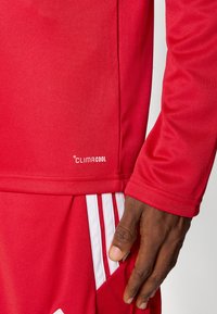 Κόκκινη αθλητική φανέλα κατασκευασμένη από αεριζόμενο ύφασμα, με το λογότυπο "CLIMACOOL", με λεία υφή και αντίθετες λευκές ρίγες στα σορτς.