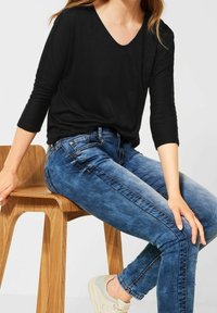 Schwarzes, locker sitzendes Oberteil mit V-Ausschnitt und Dreiviertelärmeln; kombiniert mit blauen Skinny-Jeans und beigen Sneakers, sitzt auf einem Holzstuhl.