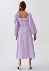 Lilac midi dress with puffy sleeves, square neckline, and pleated back detail. Smooth fabric and side slit enhance the design.