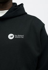 Άτομο που φοράει μαύρο φούτερ με κουκούλα με λευκό λογότυπο "New Balance Athletics Inc." στο στήθος, σε απλό φόντο.