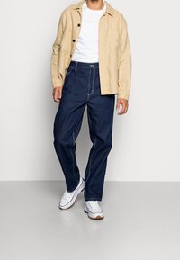 Camisa de lino beige sobre camiseta blanca, combinada con vaqueros azul oscuro. Zapatillas blancas con suelas de goma. Diseño sencillo con detalles de botones y bolsillos.