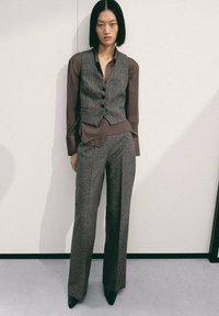 Graue Wollweste mit fünf Knöpfen, übereinander getragen über einer braunen Seidenbluse. Passende taillierte graue Hose, abgerundet mit glänzenden schwarzen Pumps.