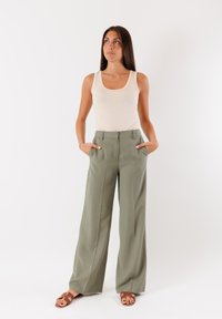 Pantaloni a gamba larga a vita alta in tessuto verde oliva, con pieghe frontali e tasche laterali, abbinati a un top beige e sandali marroni.