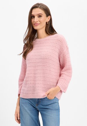 Frau mit langen braunen Haaren, die einen hellrosa gestrickten Pullover und blaue Jeans trägt, posiert mit einer Hand in der Tasche.