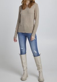 Beige Langarmpullover, figurschnitt. Zerfetzte blaue Skinny Jeans. Knielange beige Gummistiefel mit strukturiertem Sohle. Schlichte, minimalistische Stil.