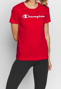 Rød bomulds T-shirt med korte ærmer, med et hvidt "Champion" logo på tværs af brystet. Blandet med sorte leggings.
