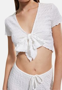 Weißes, kurzes Oberteil mit tiefem V-Ausschnitt und einer Schleifendetail an der Vorderseite. Besticht durch strukturierten, welligen Stoff. Kombiniert mit passenden weißen texturierten Bottoms.