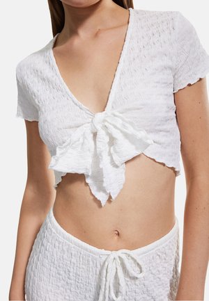 Top blanco corto con un escote en V profundo y un detalle en forma de lazo en el frente. Presenta una tela texturada y ondulada. Combinado con una parte inferior blanca texturada a juego.