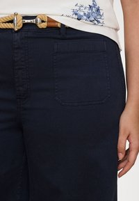 Mørk marineblå denimbukser med et klassisk design. Har en baklomme og et teksturert belte i beige med en sølvspenne.
