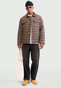 Man wearing a brown plaid quilted jacket, black pants, yellow boots, dark beanie, sunglasses, and holding a white tote bag.