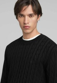 Pull en maille torsadée noire avec col rond et motif texturé. Détail subtil du logo sur la poitrine. Tissu doux avec poignets côtelés.