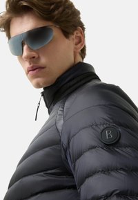 Czarna kurtka puchowa z wysokim kołnierzem i pikowaną strukturą. Na lewym ramieniu znajduje się okrągłe, wytłaczane logo, a z przodu zamek błyskawiczny.