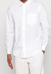 Man in een wit overhemden met lange mouwen en een borstzak, gedragen met witte broek, met één hand in de zak.