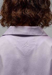 Camicia viola chiaro con colletto, caratterizzata da un design testurizzato. Un logo Jumpman ricamato di colore bianco si trova sul retro, sotto il colletto.
