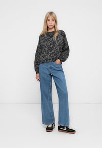 Donna in piedi con indosso un maglione grigio a stampa leopardata, jeans a gamba larga blu e sneaker nere con suola bianca, su uno sfondo bianco semplice.